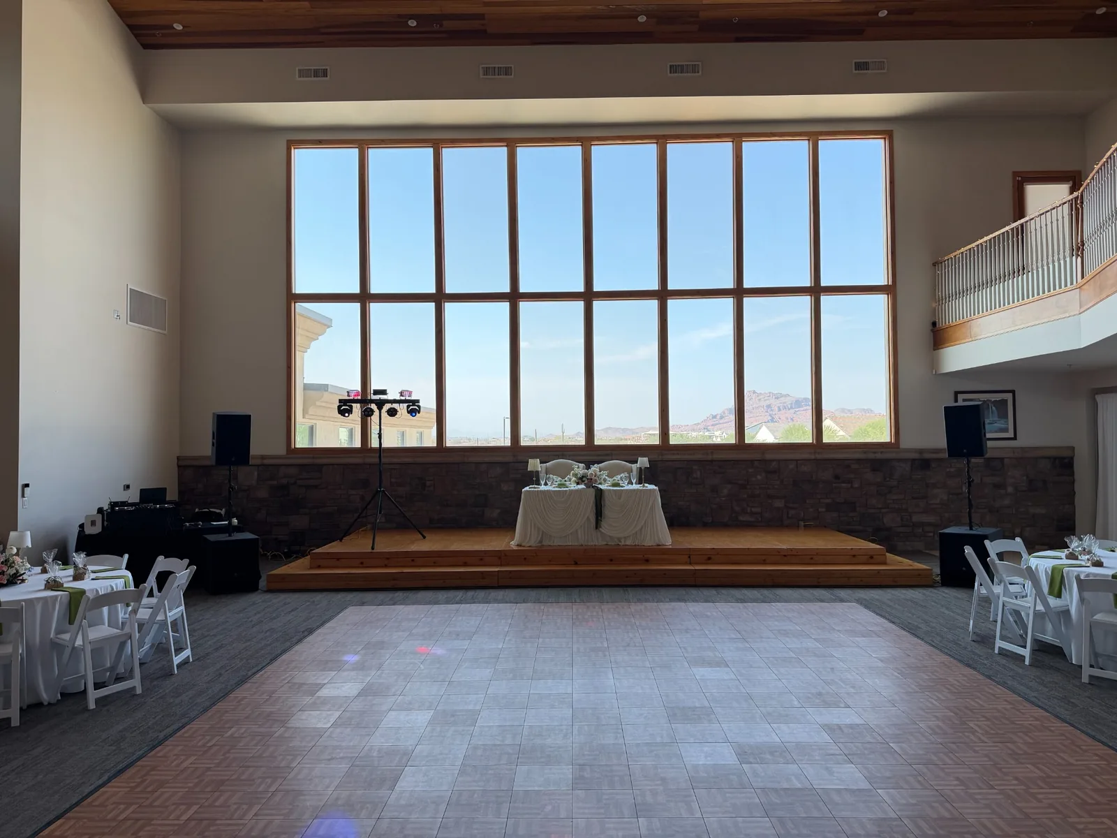 Empty reception hall with a wood dance floor, sweetheart table, and DJ speakers and lights flanking the stage
